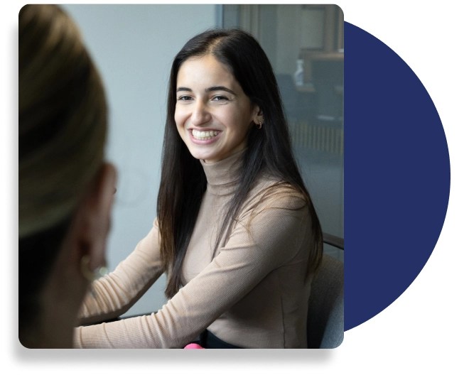 Young woman with long dark hair in a light brown turtleneck, smiling during a conversation in a modern office setting with glass partitions and a blurred background.