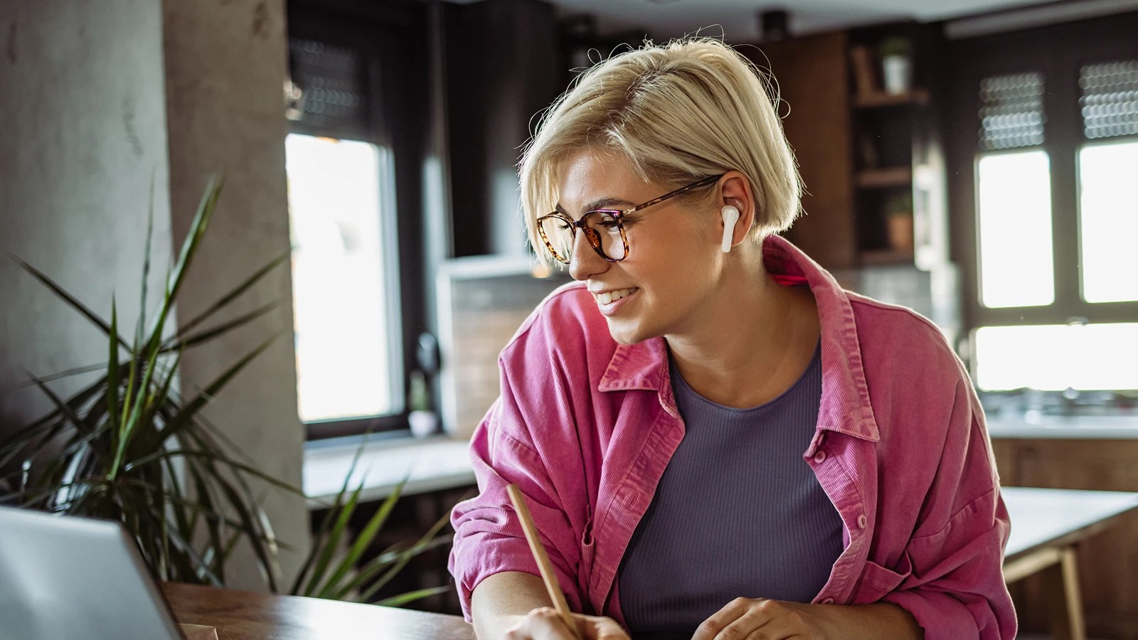 Smiling person with short blonde hair, glasses, and white earbuds, writing with a pencil while looking at a laptop in a cozy kitchen space with plants and large windows.