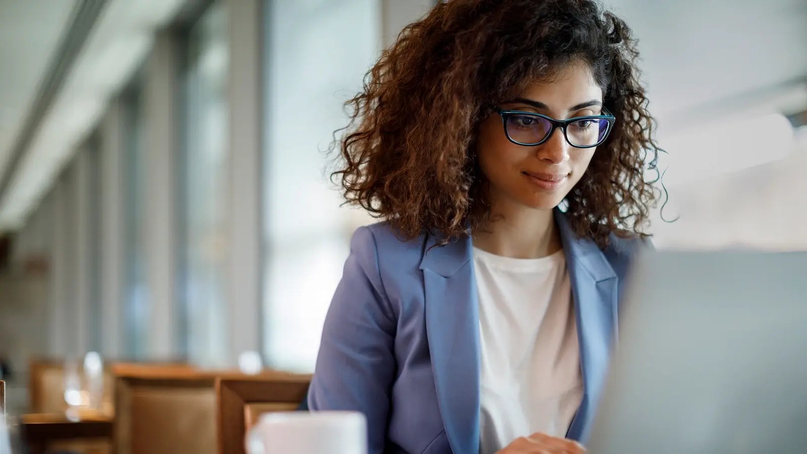 Person with curly hair and glasses, wearing a light blue blazer and white top, working on a laptop in a bright café or office space with large windows and a coffee cup nearby.