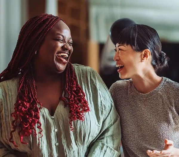 Two people sharing a laugh indoors, one with long red braids in a light green shirt, the other with short dark hair in a gray top, standing close together in a warm, casual setting with a large plant in the background.