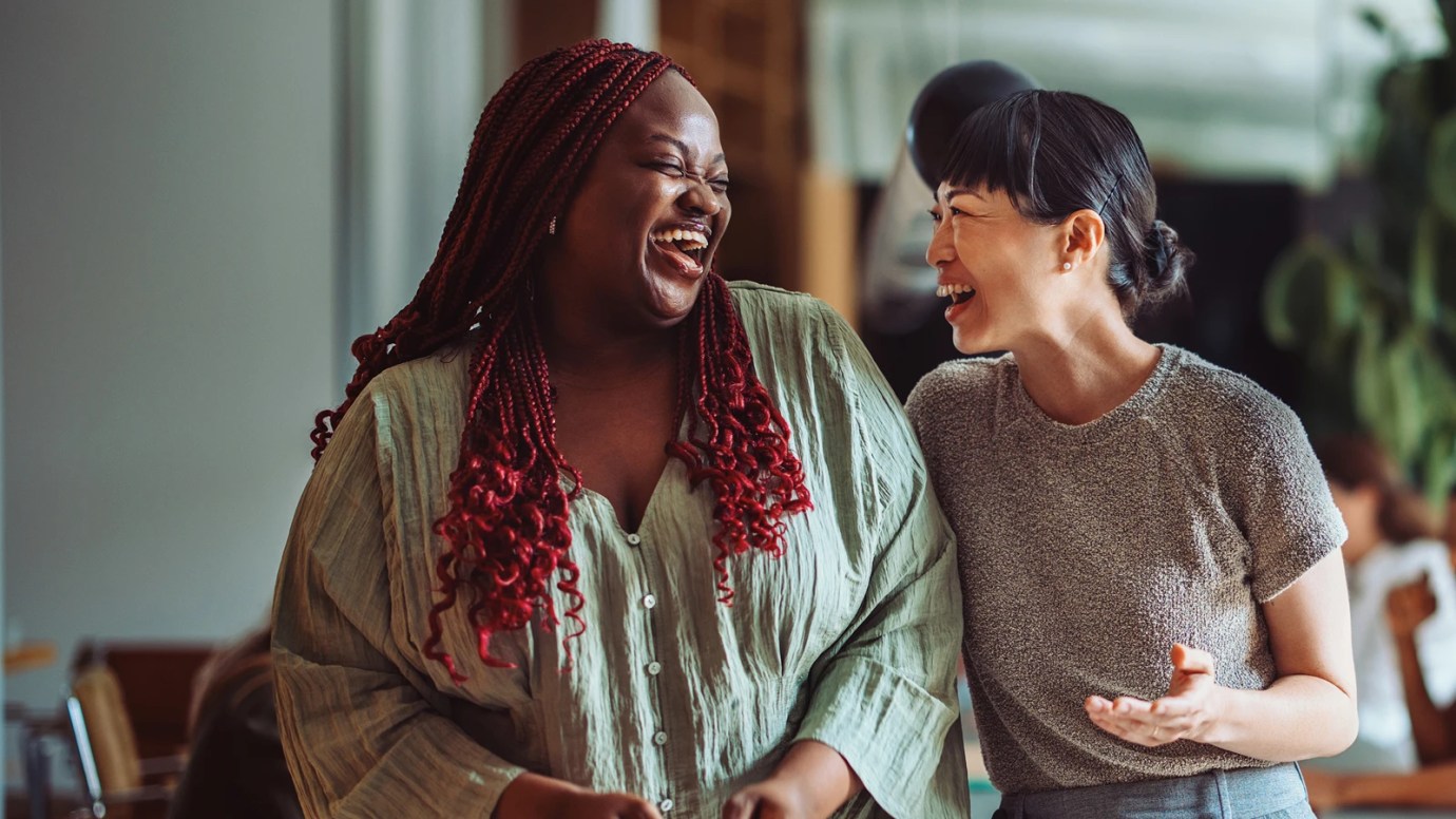 Two people sharing a laugh indoors, one with long red braids in a light green shirt, the other with short dark hair in a gray top, standing close together in a warm, casual setting with a large plant in the background.