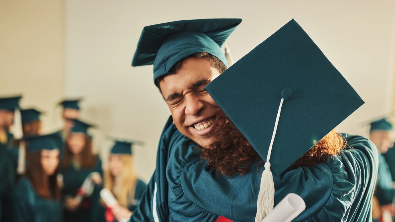 Two graduates in caps and gowns embracing joyfully indoors, one smiling with eyes closed and the other holding a diploma, surrounded by fellow graduates celebrating.