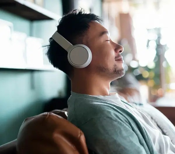 Person relaxing on a couch with eyes closed, wearing over-ear headphones and a teal cardigan, surrounded by warm decor and natural light in a peaceful indoor setting.
