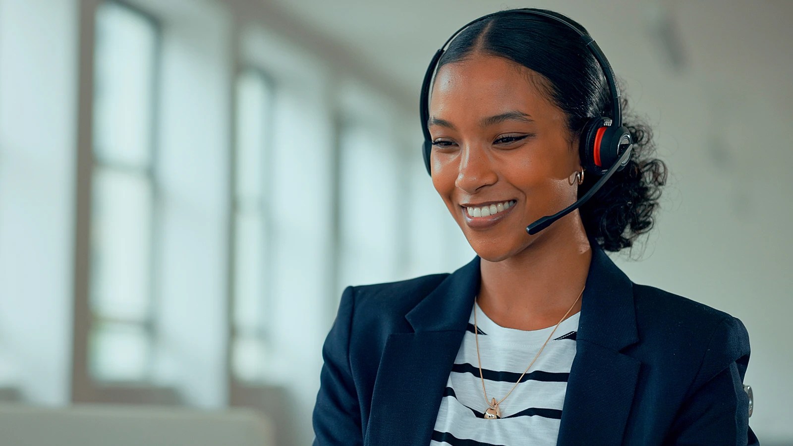 Smiling person wearing a headset and striped shirt under a dark blazer, seated in a bright office space with large windows, suggesting a professional or customer service setting.