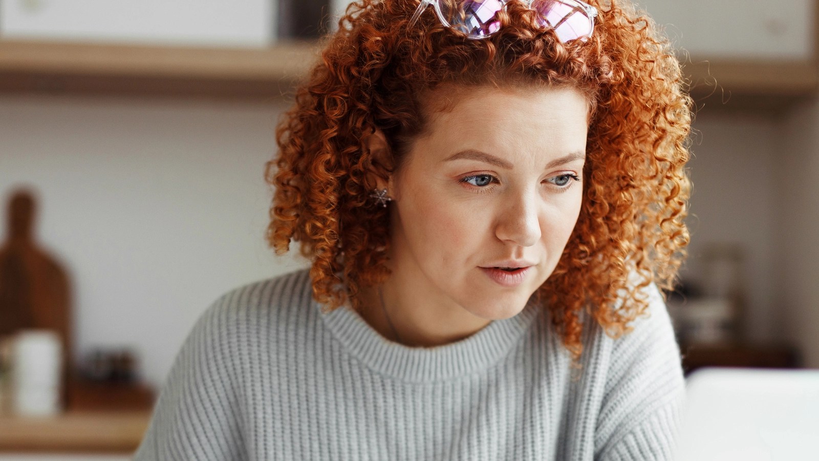 Person with curly red hair wearing a gray sweater and sunglasses on their head, focused on something off-screen in a cozy room with shelves and kitchen items.