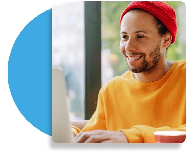 Person smiling while working on a laptop, wearing a red beanie and yellow sweatshirt, seated indoors near a window with greenery outside and a red cup on the table.