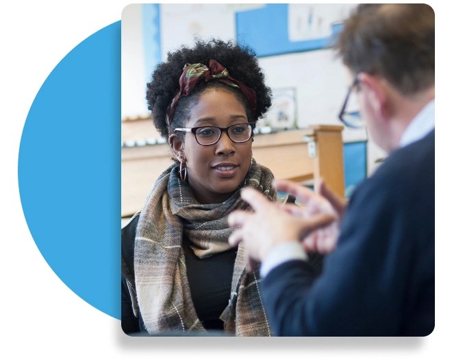 Two people engaged in conversation indoors, one gesturing with both hands while the other, wearing glasses, a patterned scarf, and headband, listens attentively in a space with bookshelves and educational posters.