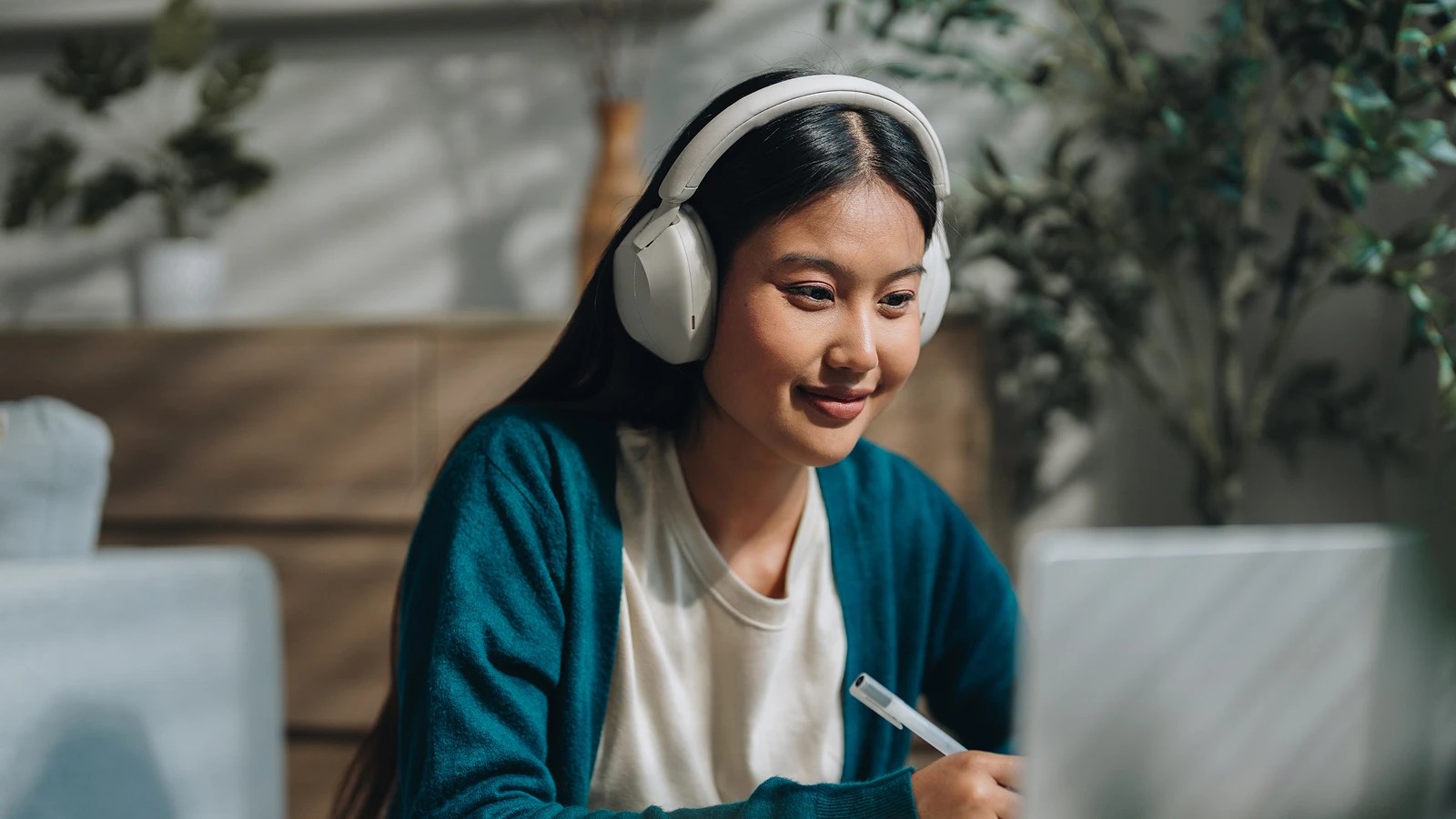 Person in a teal cardigan and over-ear headphones, focused on a laptop while holding a pen, seated in a cozy indoor space with plants and soft lighting.
