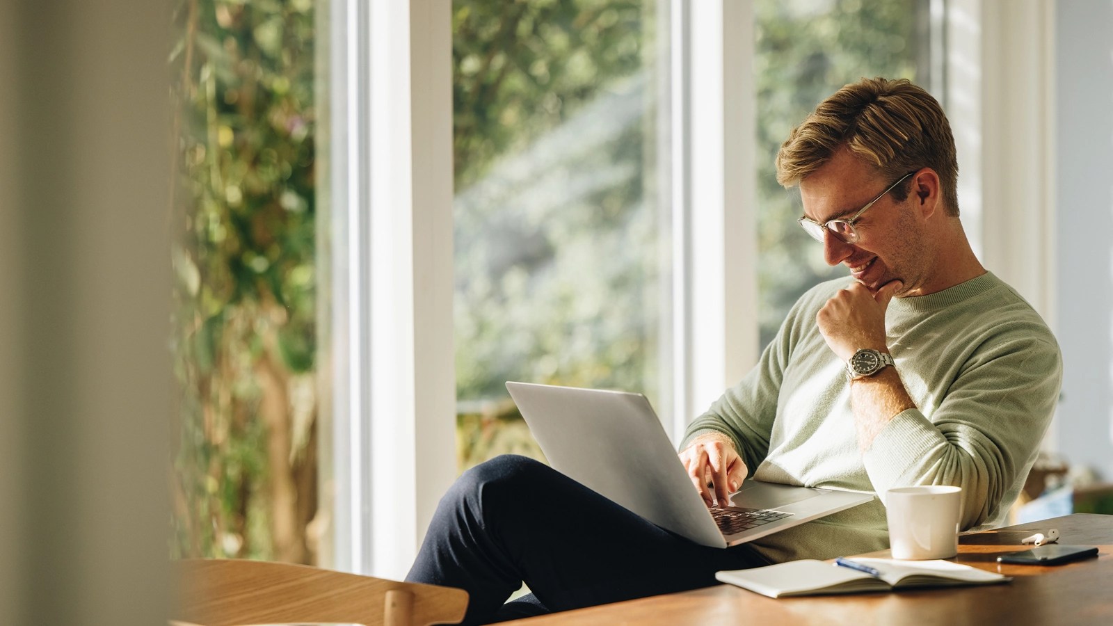 Person in a light sweater and glasses working on a laptop near large windows, with a coffee cup, notebook, and glasses on the table, in a bright indoor space overlooking greenery.