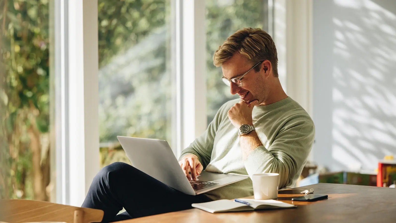 Person in a light sweater and glasses working on a laptop near large windows, with a coffee cup, notebook, and glasses on the table, in a bright indoor space overlooking greenery.