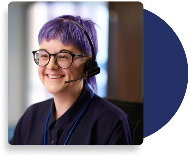 Smiling person with vibrant purple hair, glasses, and a headset, wearing a navy collared shirt and lanyard in a modern, softly blurred indoor setting.
