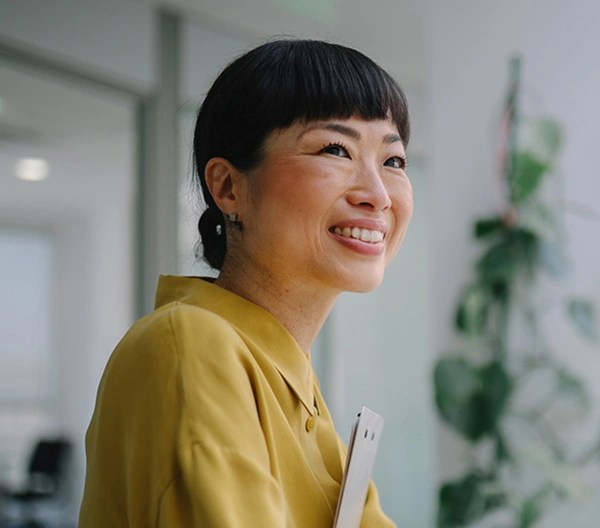 Person in a mustard yellow blouse smiling and holding a tablet in a modern office with glass doors, furniture, and a tall plant.
