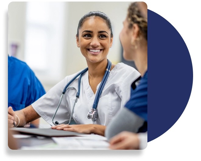 A healthcare professional, wearing hospital scrubs and a stethoscope around the neck, smiles with another person.