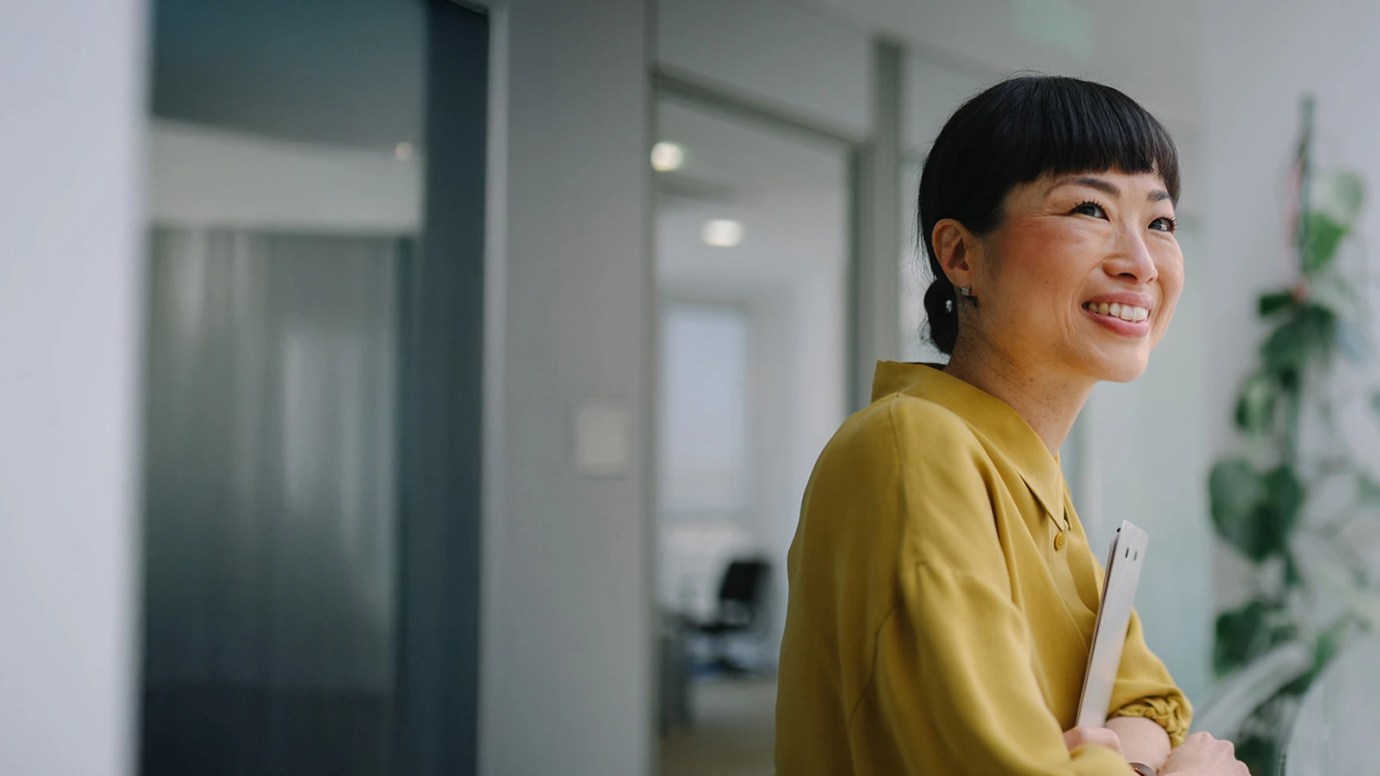 Person in a mustard yellow blouse smiling and holding a tablet in a modern office with glass doors, furniture, and a tall plant.