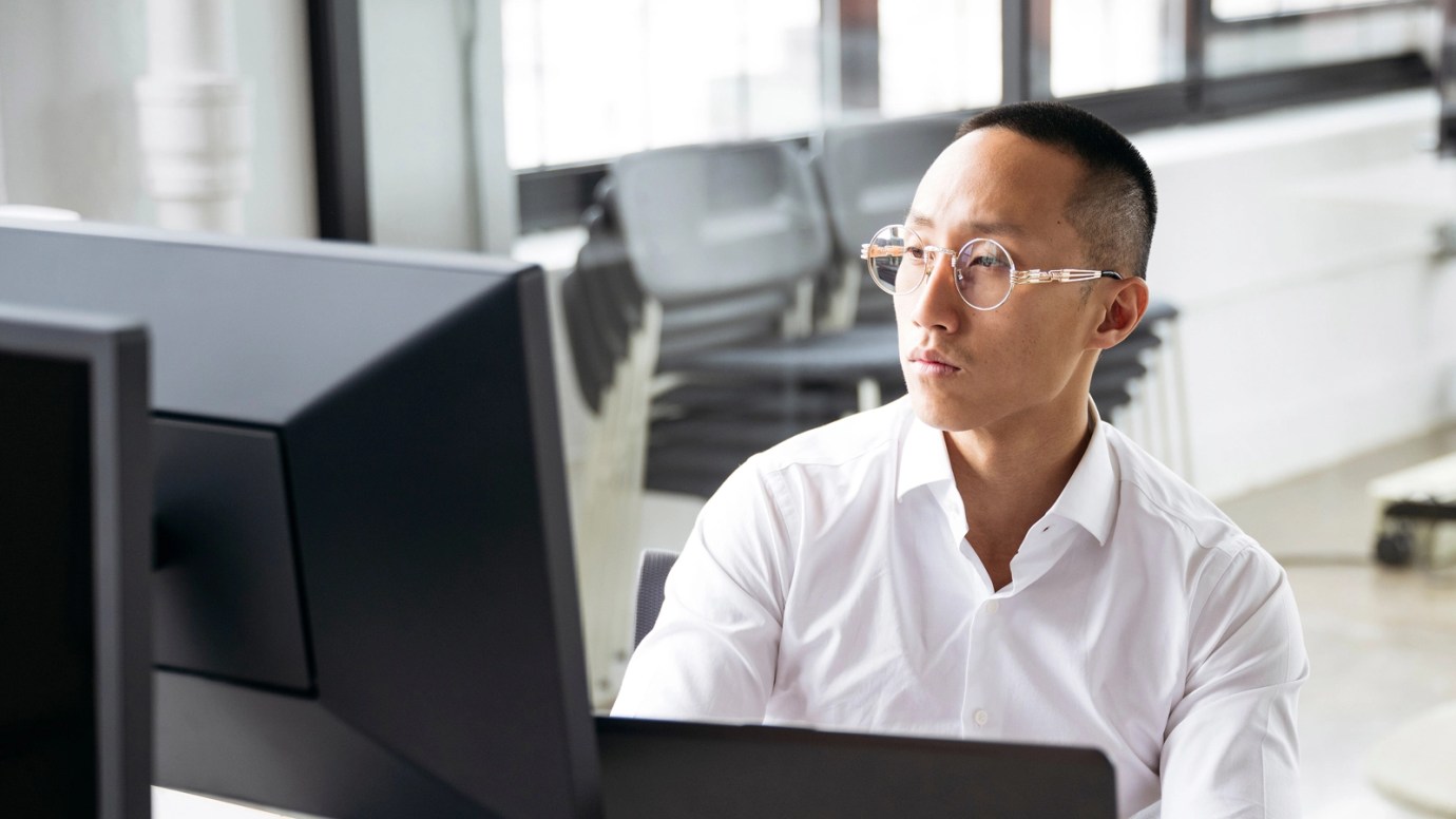 An individual, wearing a white blouse and glasses, sits at their computer working.