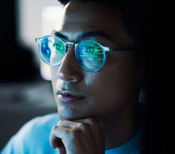Close-up of a person wearing glasses, thoughtfully gazing at a computer screen with code reflected in the lenses.