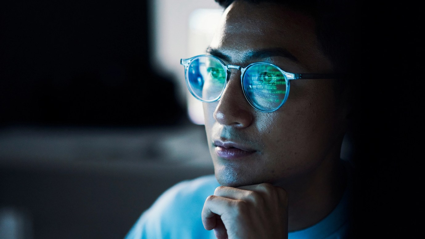 Close-up of a person wearing glasses, thoughtfully gazing at a computer screen with code reflected in the lenses.