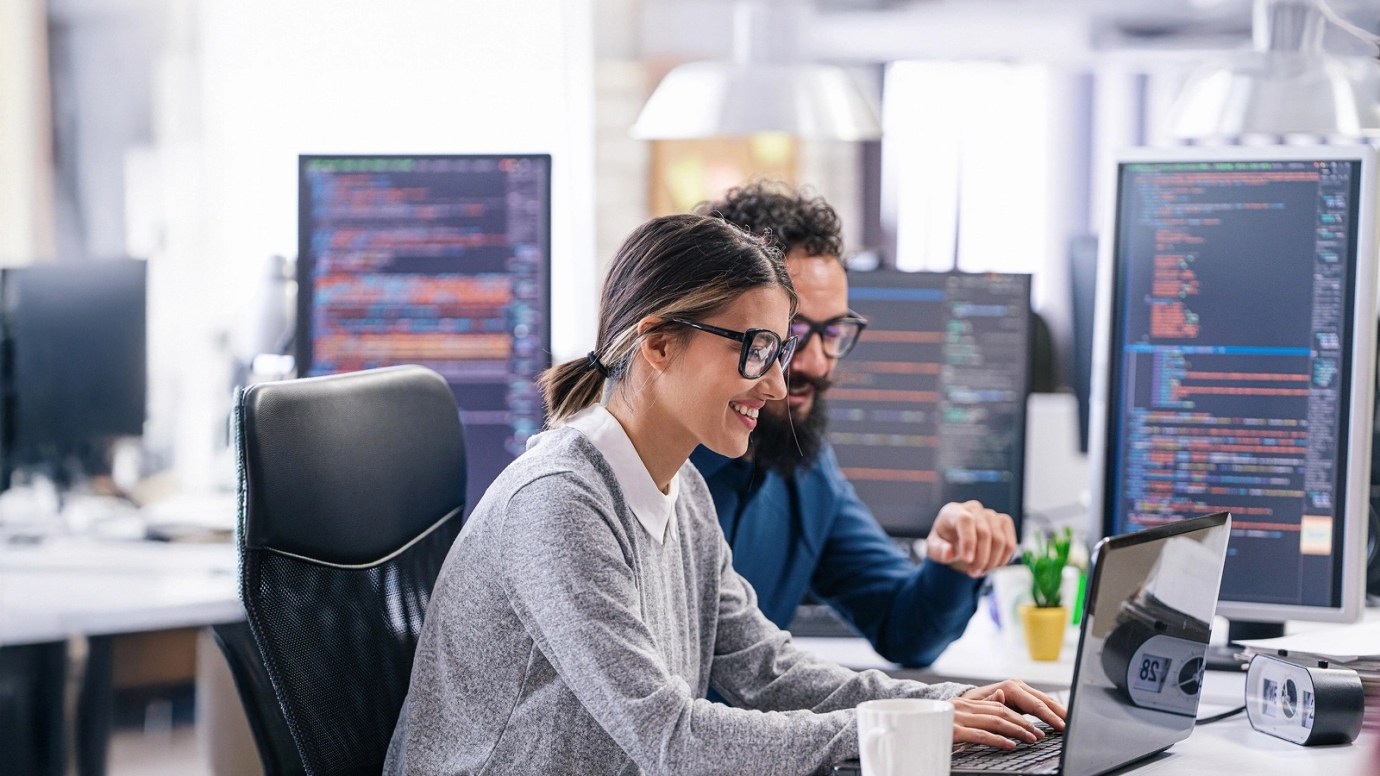 Two people working together at a desk in a tech office, focused on a laptop and surrounded by monitors displaying code.