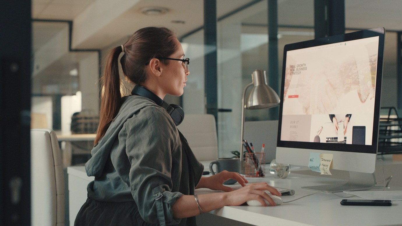 An individual with brown tied back hair, wearing a grey blouse and glasses, sits at their desk, hand on a mouse, in front a desktop computer.