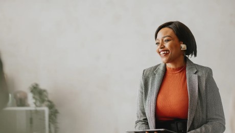 A professional, wearing an orange shirt and grey jacket, smiles sitting on a desk.