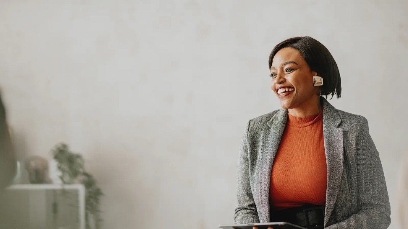 A professional, wearing an orange shirt and grey jacket, smiles sitting on a desk.