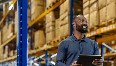 Person holding a tablet and inspecting shelves stacked with cardboard boxes in a brightly lit warehouse.