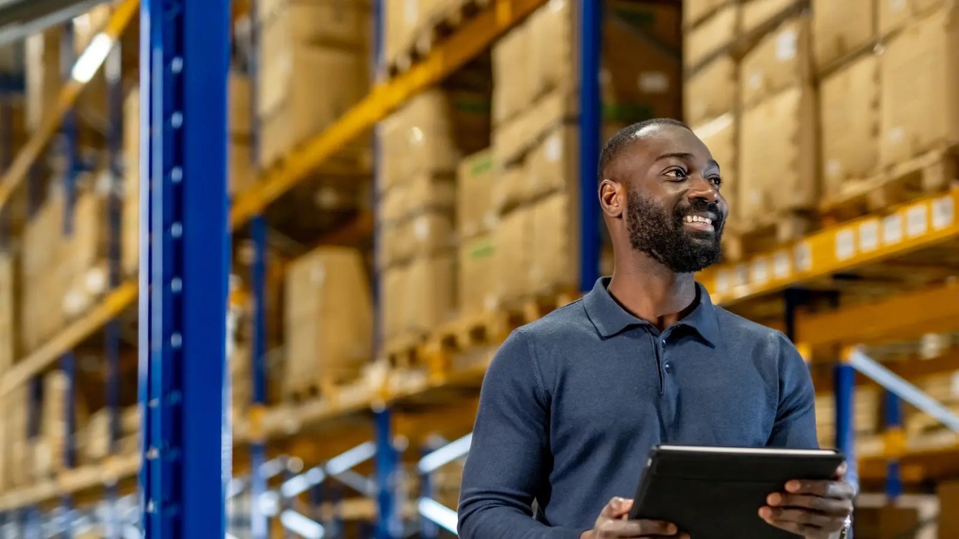 Person holding a tablet and inspecting shelves stacked with cardboard boxes in a brightly lit warehouse.