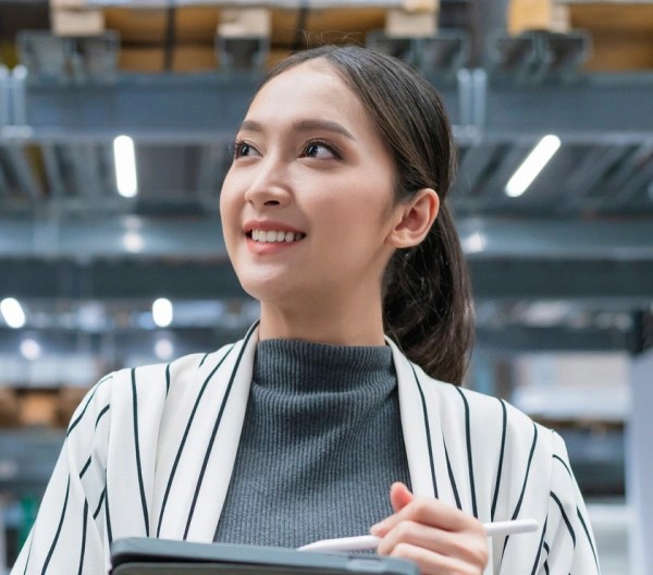Person in a striped outer garment holding a clipboard and pen, looking upward in a bright warehouse with tall shelves and organized boxes—suggesting inventory or inspection work.