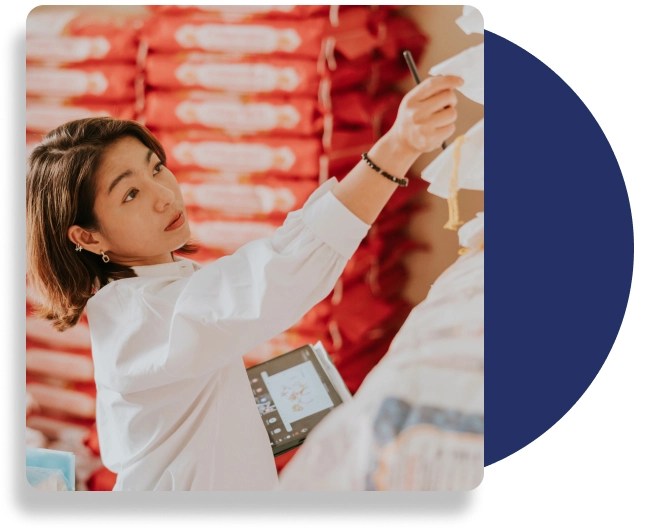 A female professional wearing a white blouse inspects products in a warehouse, while holding a digital tablet.