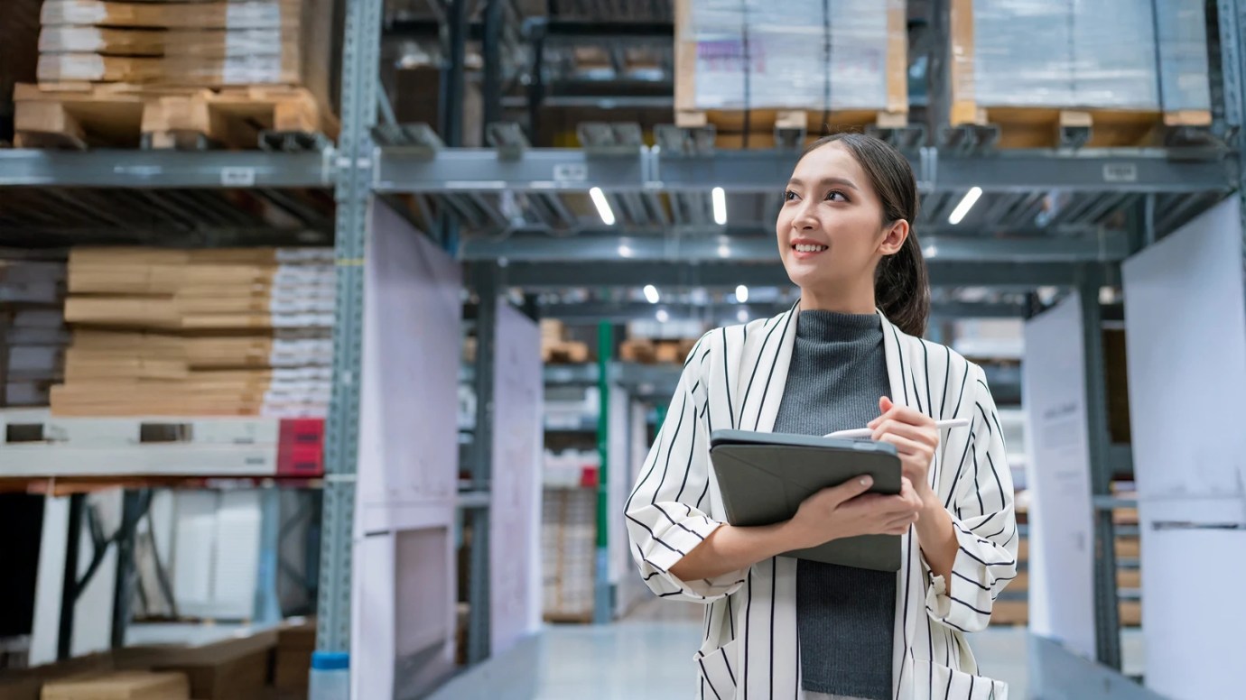 Person in a striped outer garment holding a clipboard and pen, looking upward in a bright warehouse with tall shelves and organized boxes—suggesting inventory or inspection work.