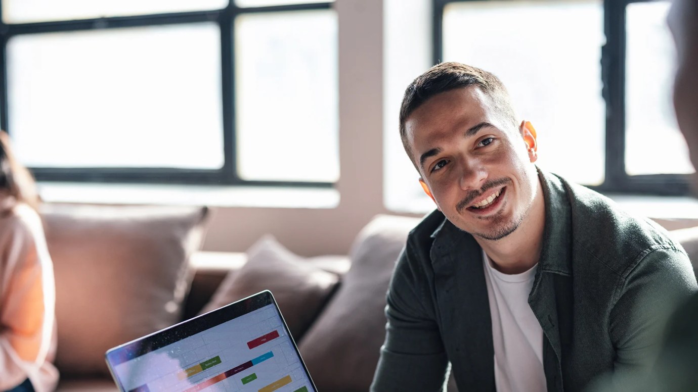 A smiling person displaying their laptop to someone in an indoor setting.