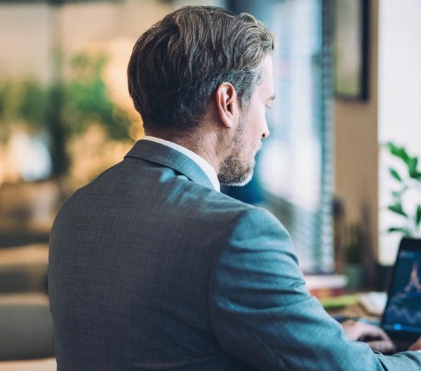 Person in a suit working on a laptop displaying candlestick charts, seated in a bright office with plants.