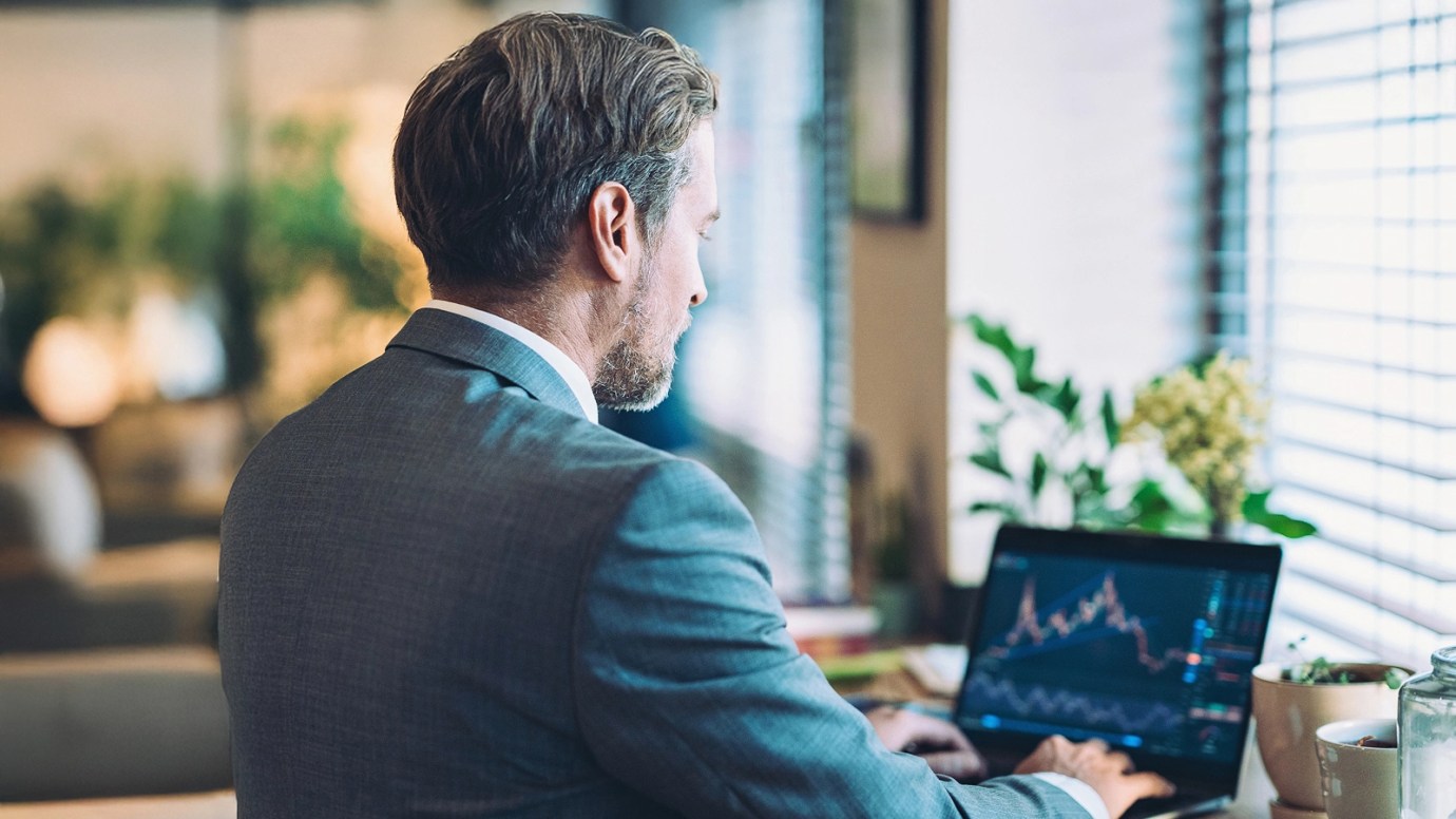 Person in a suit working on a laptop displaying candlestick charts, seated in a bright office with plants.