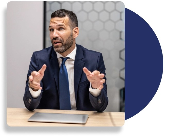 An individual wearing a dark blue tie, white dress shirt and dark blue suit jacket, explains, whilst sitting at a desk, with a closed laptop.