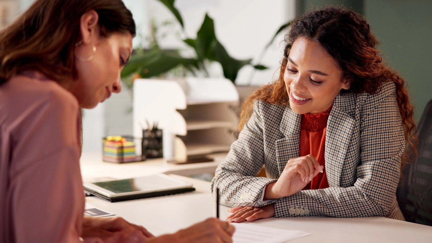 Two people seated at a desk in an office, one writing on paper while the other smiles and observes,