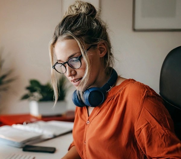 A female professional wearing an orange blouse writes on a small whiteboard, sitting at a desk in front of a desktop computer.