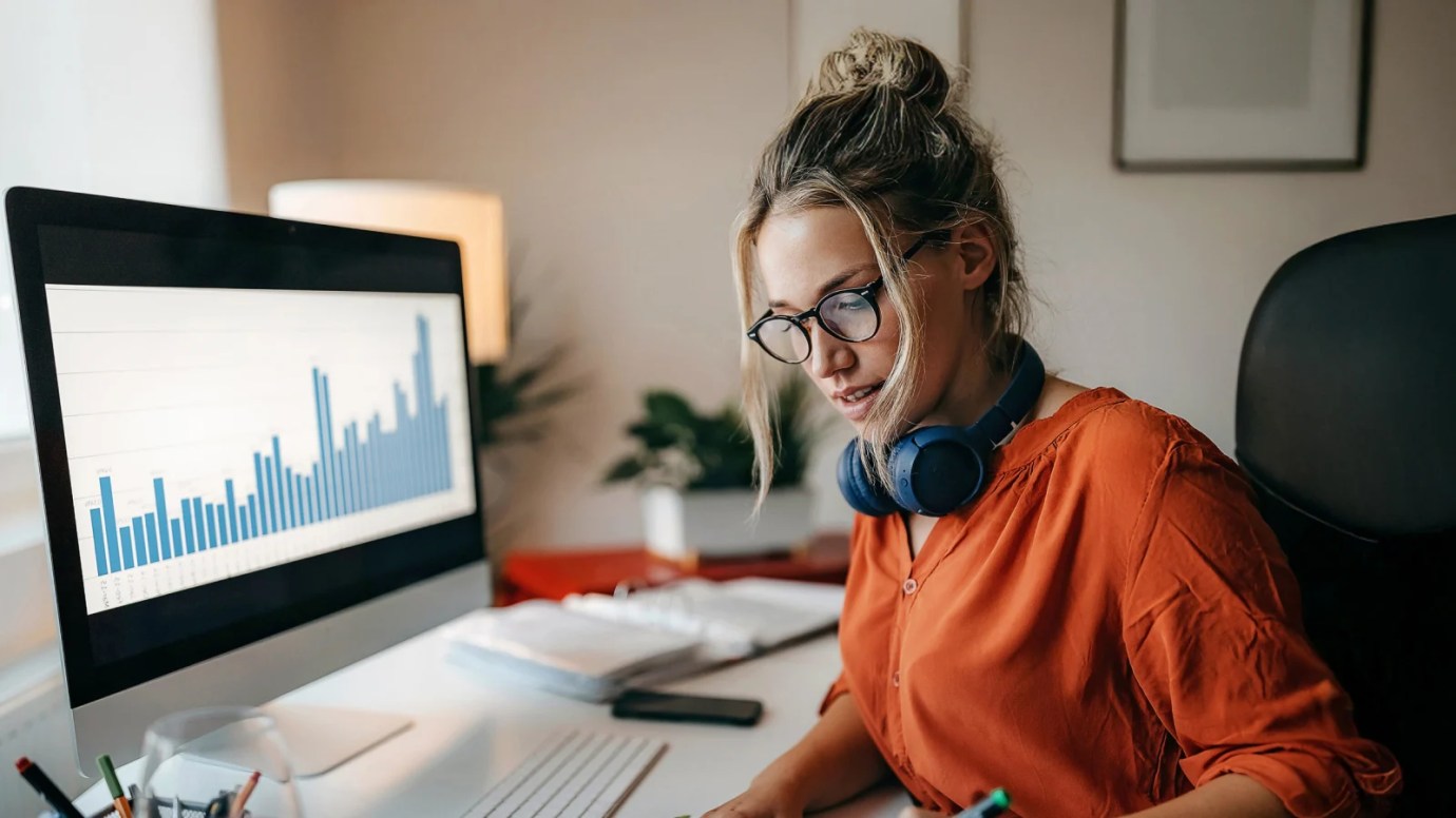 A female professional wearing an orange blouse writes on a small whiteboard, sitting at a desk in front of a desktop computer.
