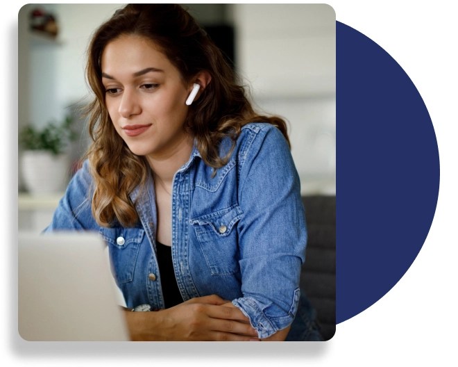 A woman sits at her laptop, wearing wireless earphones, wearing a blue blouse.