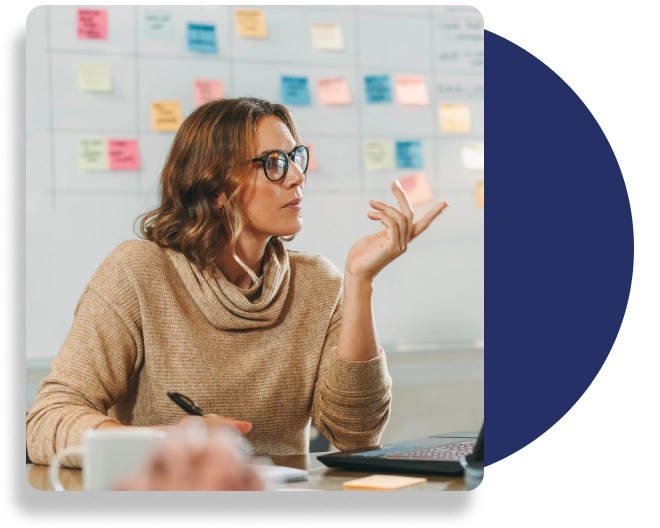 A professional wearing a beige jumper, takes notes and communicate, sitting in front a wall with sticky notes on it.