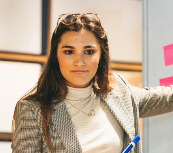 An individual wearing a white shirt, jacket, and jeans presents sticky notes to other colleagues in an indoor setting.