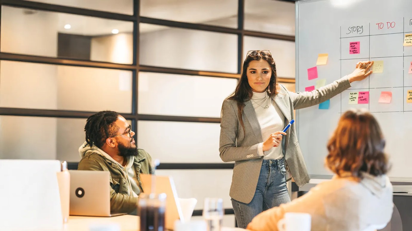 An individual wearing a white shirt, jacket, and jeans presents sticky notes to other colleagues in an indoor setting.
