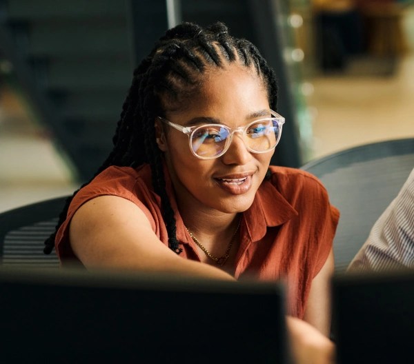 An individual wearing glasses and a short sleeved light brown shirt, points to something on a computer screen, sitting next to another individual.