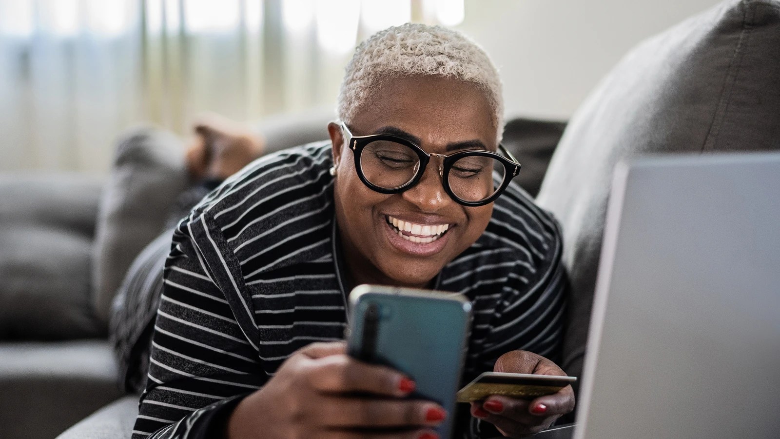 Person lying on a couch, smiling at a smartphone while holding a credit card, with a laptop nearby in a softly lit room.