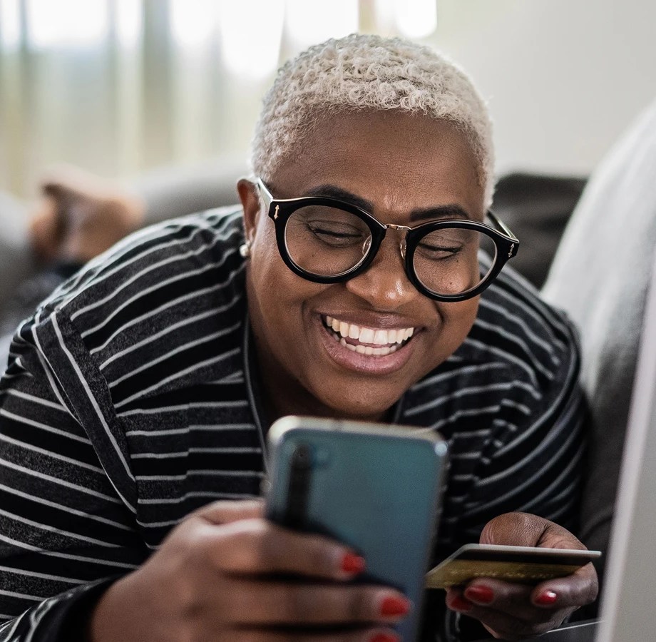 Person lying on a couch, smiling at a smartphone while holding a credit card, with a laptop nearby in a softly lit room.