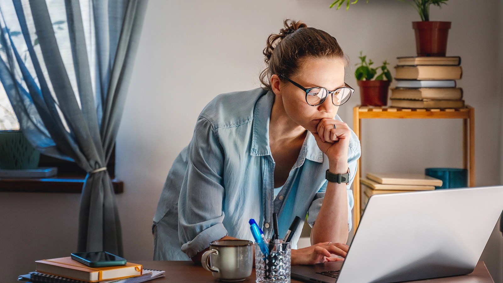 Person in glasses and a light blue shirt working at a desk with a laptop, surrounded by study items in a softly lit home office with books and a plant in the background.