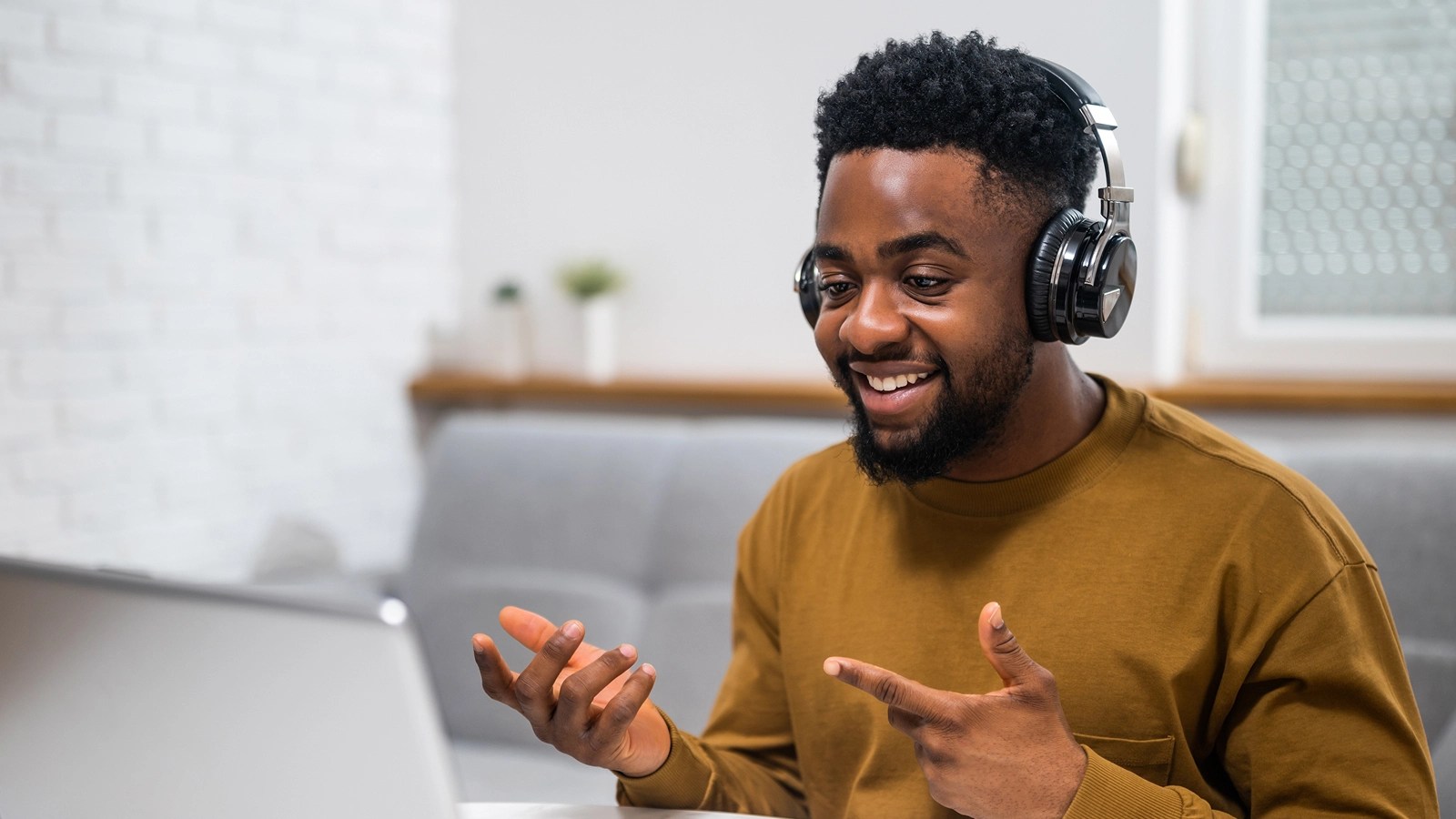 Person in a brown shirt and headphones smiling and gesturing during a video call, seated at a laptop in a cozy home setting with a couch and white brick wall.