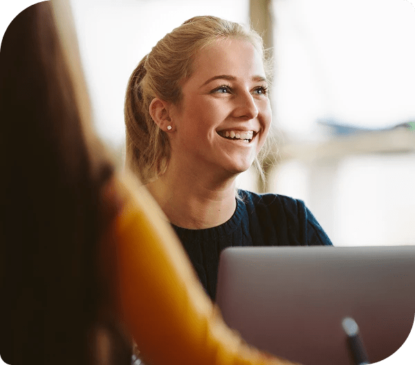 Smiling person with light hair pulled back, seated at a laptop in a softly lit indoor space, engaged in conversation with another individual.