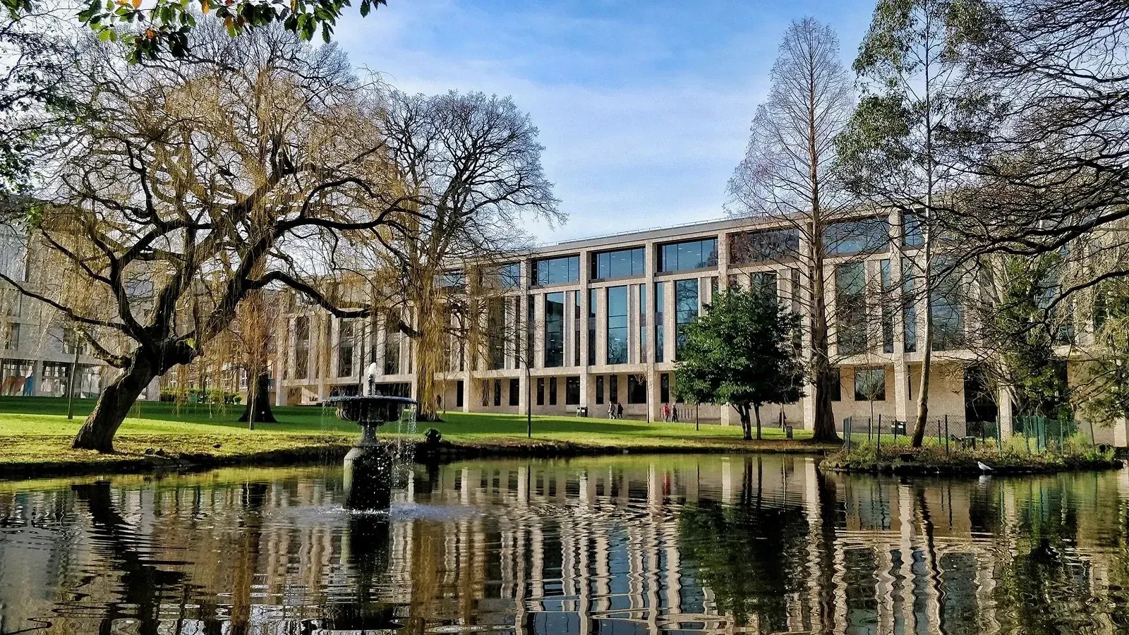 Modern glass-fronted library at Roehampton University, reflected in a pond with a central fountain, surrounded by trees and landscaped greenery on a clear day.