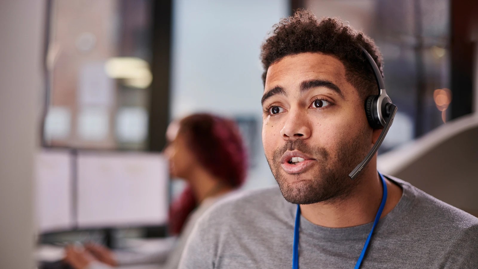 Person wearing a headset, engaged in conversation in a bright office setting, with another individual blurred in the background seated at computer monitors.