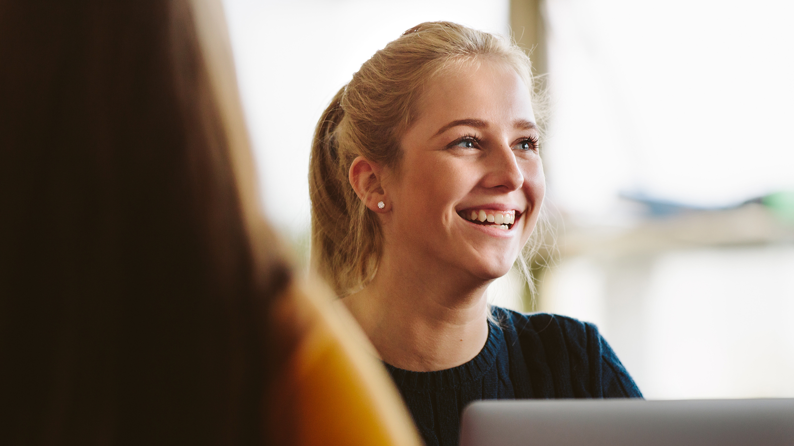 Smiling person with light hair pulled back, seated at a laptop in a softly lit indoor space, engaged in conversation with another individual.